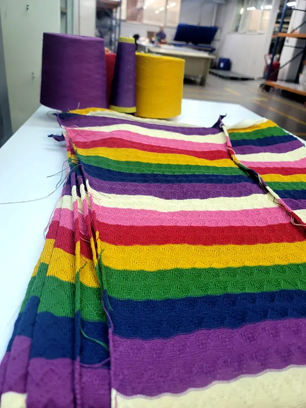 Rainbow striped knit panels on a table with purple and yellow yarn cones