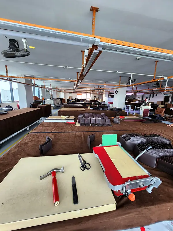 Leather workbench with a red hammer, a knife, scissors and a small edge press, the factory floor behind