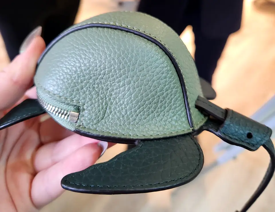 A hand holding a sage and dark green pebbled-leather pouch shaped like a sea turtle