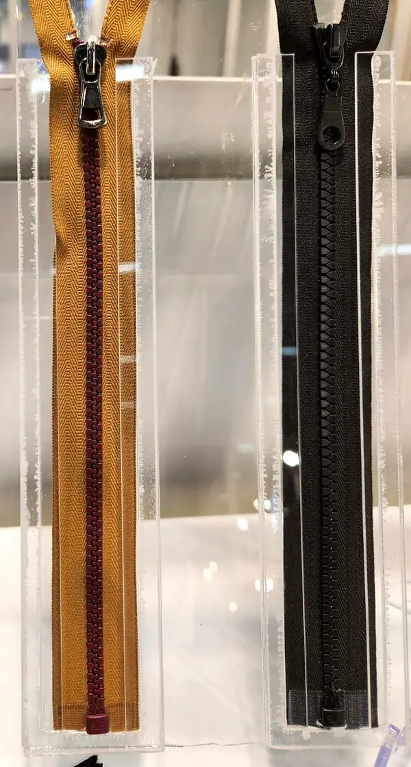A tan zip with burgundy teeth next to a chunky black zip in a clear display stand